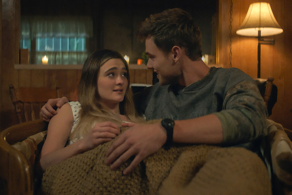 A young couple cuddles on a couch under a brown blanket in a cozy, warmly-lit living room with wooden walls, candles, and a lamp creating a relaxed, intimate atmosphere.