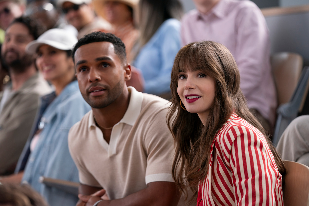 Lucien Laviscount as Alfie and Lily Collins as Emily sit at a stadium together in Season 4 of ‘Emily in Paris.’