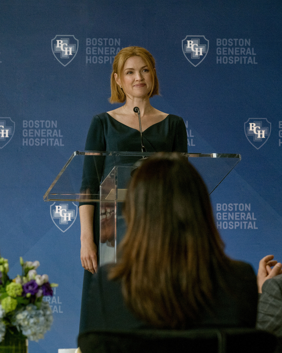 Woman speaking at podium at Boston General Hospital event, audience seated, large poster to the right shows smiling boy and “Donate Today!” message, bouquet of flowers on table, formal indoor setting.