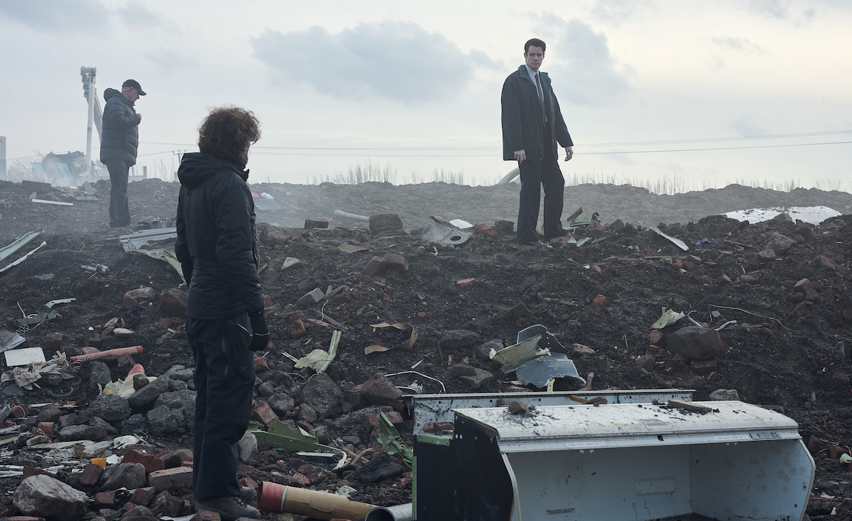 Three people inspecting debris and rubble on a desolate, cloudy landscape; two stand among scattered wreckage and broken objects, while one observes from a distance, suggesting a post-disaster or crime scene investigation setting.