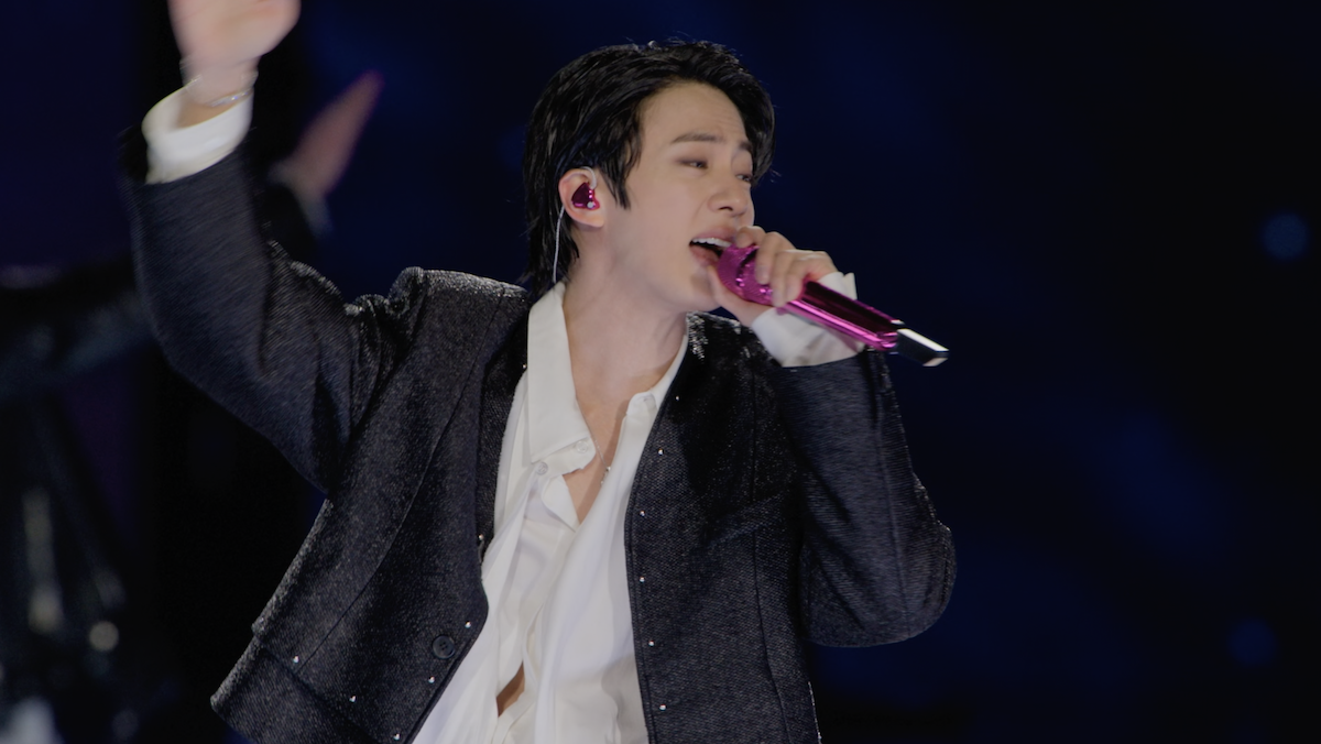 Singer with dark hair performs on stage wearing a black jacket and white shirt, holding a pink microphone, with blurred audience and stage lighting in the background, capturing an energetic concert atmosphere.