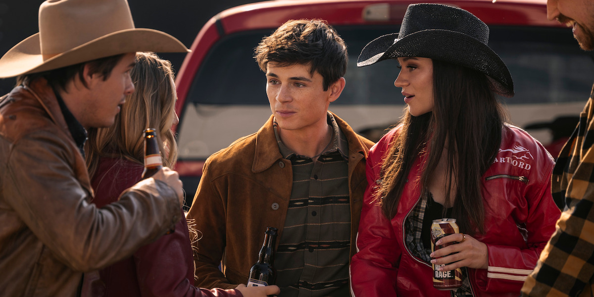 Group of young adults wearing cowboy hats and casual jackets socialize outdoors by the tailgate of a red truck, holding bottled drinks, in a relaxed daylight setting.