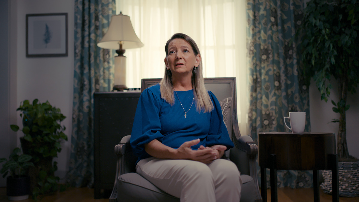 Woman in blue shirt sitting in an armchair, speaking, in a softly lit living room with plants, a mug on a side table, framed art, a lamp, and floral curtains in the background.