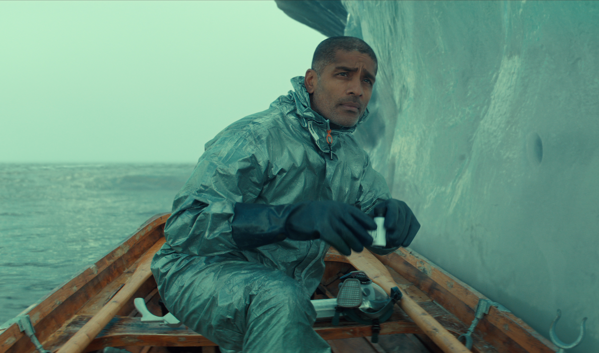 A man in waterproof gear sits in a wooden boat beside a large iceberg on a cold, overcast day, holding a small object and appearing thoughtful as he looks ahead, with the icy ocean in the background.