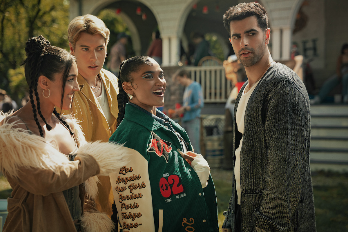 Four young adults stand together outside at a social gathering, with a house and other people in the background, appearing to converse and enjoy a lively daytime event.