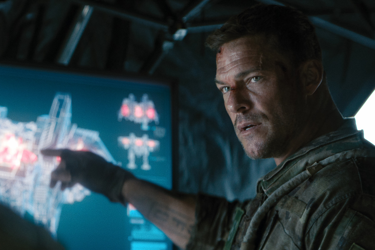 A man in military gear points at a digital tactical map on a screen inside a dimly lit command tent, suggesting a strategic or tense planning environment.