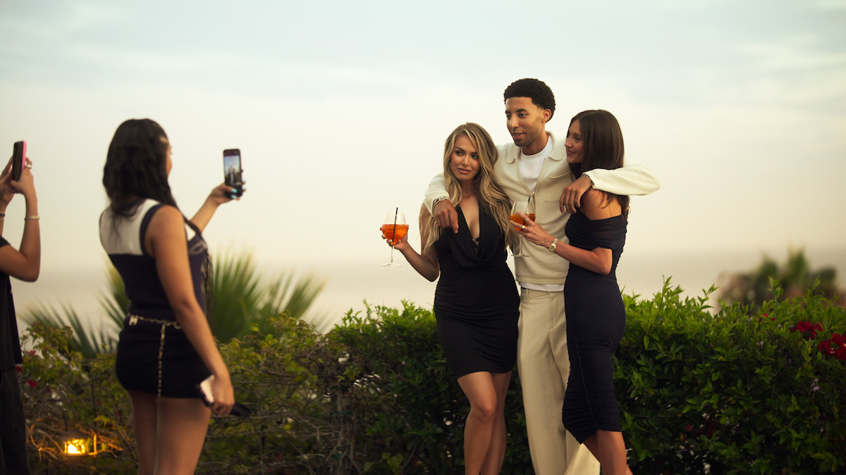 Three people posing together outdoors near greenery at sunset with drinks in hand while another person takes their photo using a smartphone. The setting appears to be a garden or terrace with a scenic view.