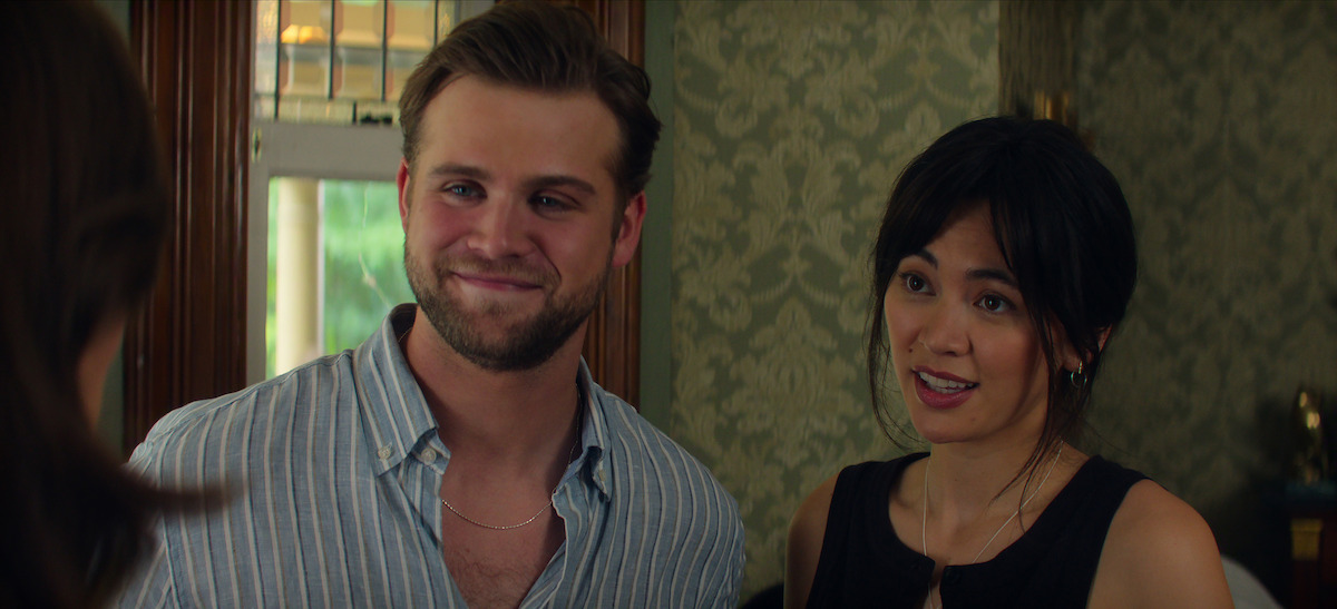 Leo Woodall, who plays Vladimir, and Jessica Henwick, who plays Cynthia, stand indoors, smiling and talking to another person, with patterned wallpaper and windows in the background, creating a casual atmosphere.