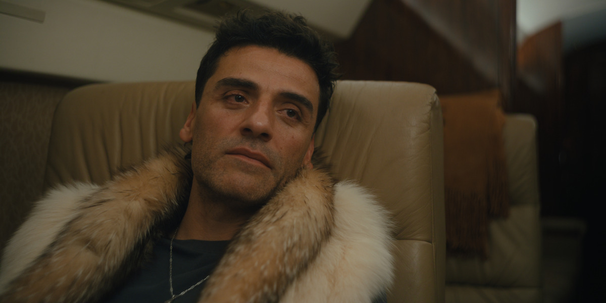 Man wearing a fur-collared coat sits in a luxurious private jet, looking pensive and relaxed against a beige leather seat, with dim lighting and wood-paneled walls in the background.