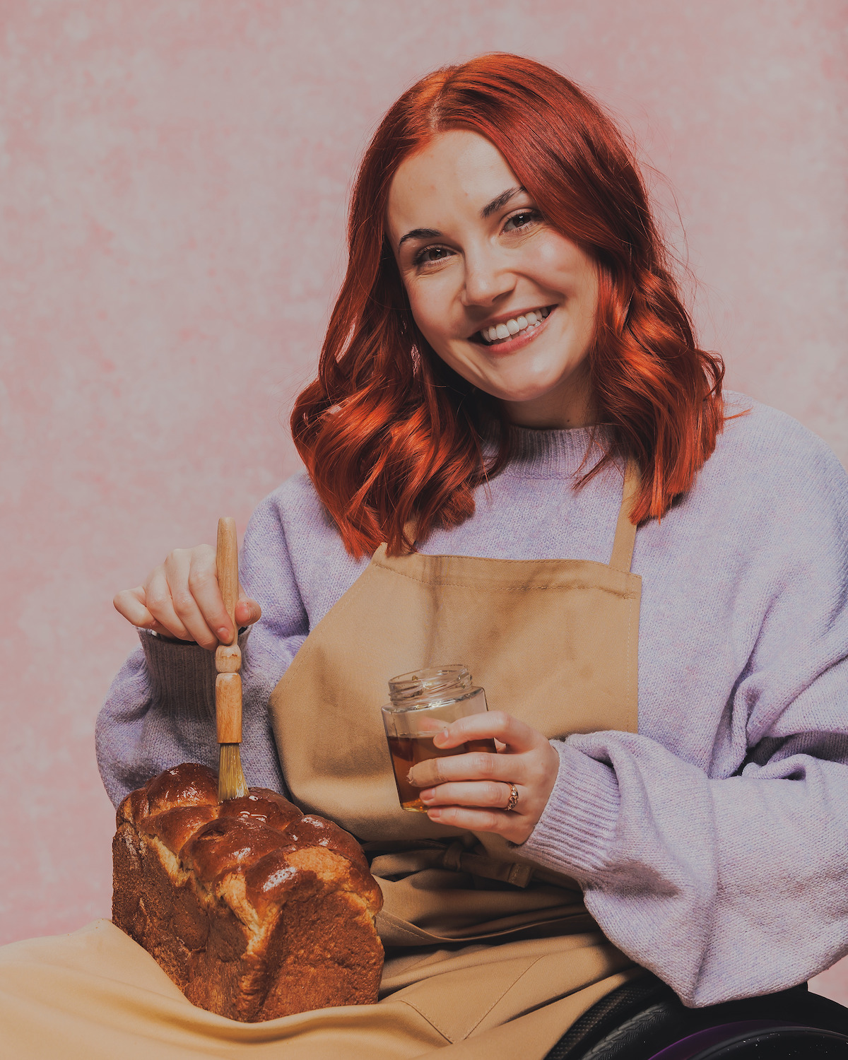 Smiling woman with red hair, wearing an apron and lavender sweater, seated indoors against a pink background, brushing glaze onto a loaf of bread while holding a jar.