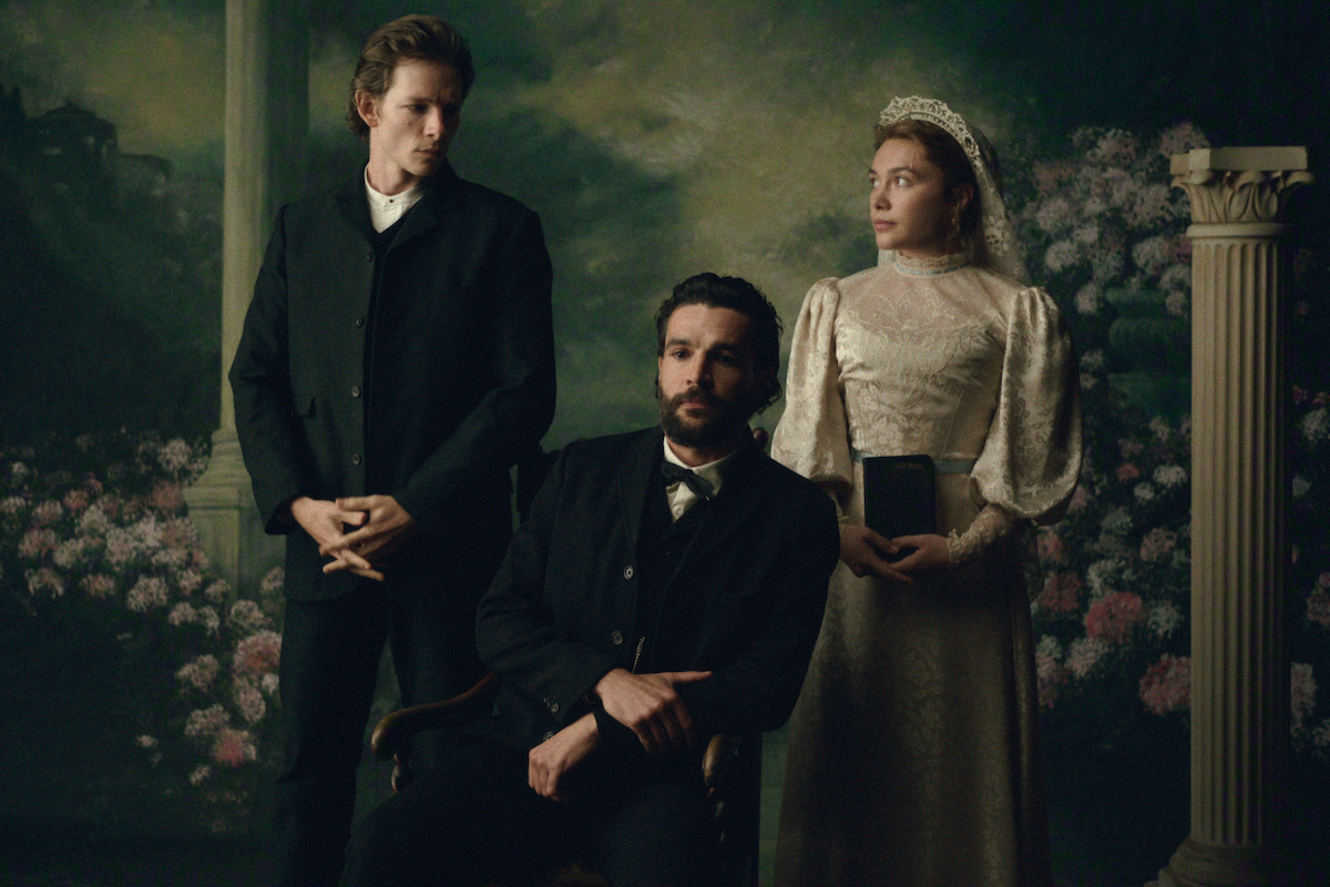 Three people in Victorian-era clothing pose in a formal portrait setting with floral and classical column background; one man seated, one man standing, and a woman in a white dress holding a book.