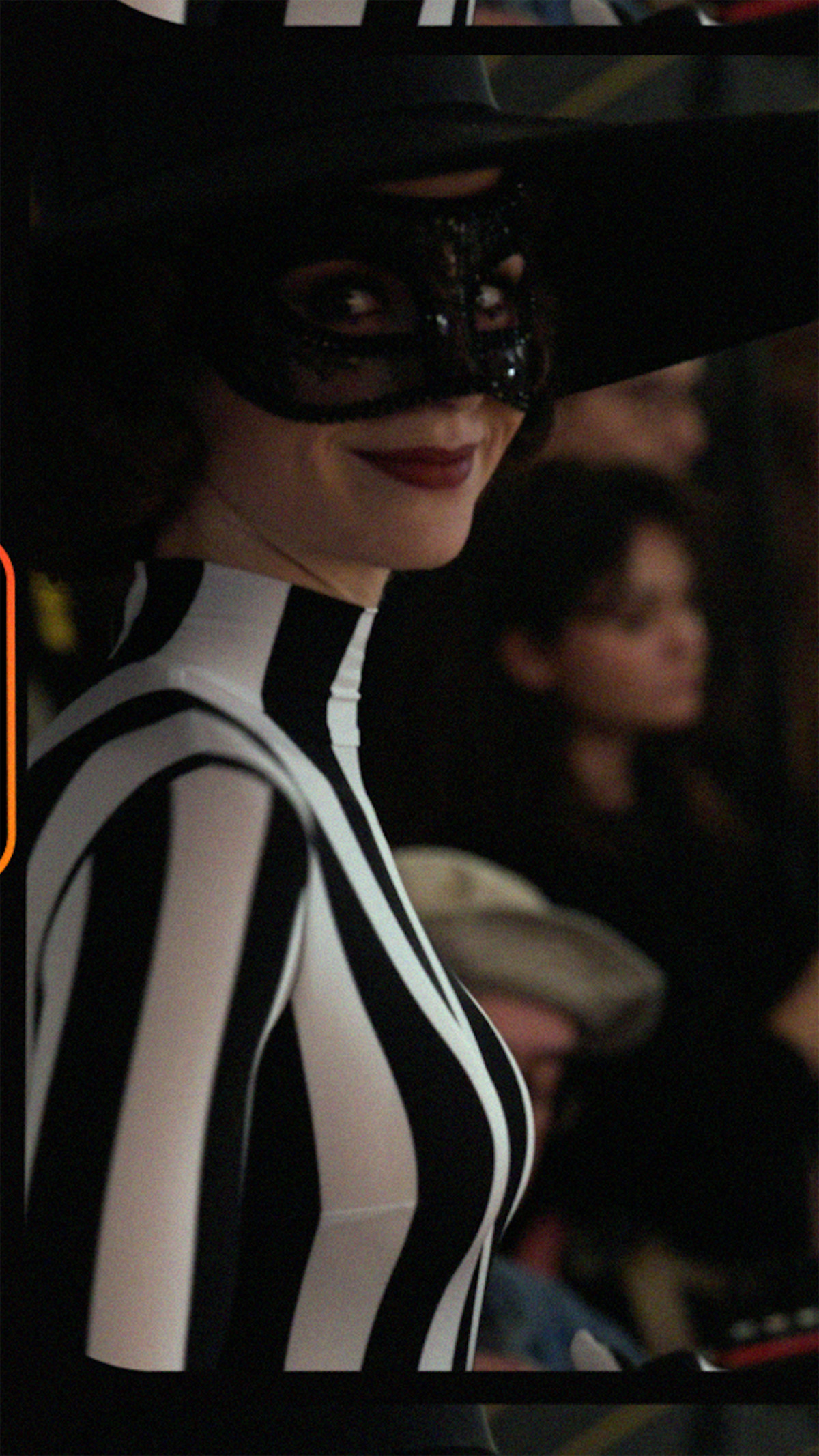 Person in a black and white striped costume, wearing a black mask and large hat, smiling at an indoor event with a blurred audience in the background.