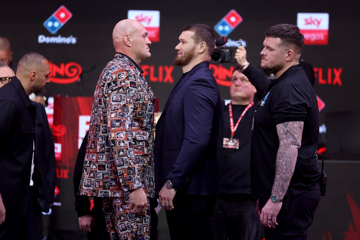 Two men facing off at a press event, one in a patterned suit and the other in a dark suit, surrounded by security and media, with event branding and sponsors visible in the background.