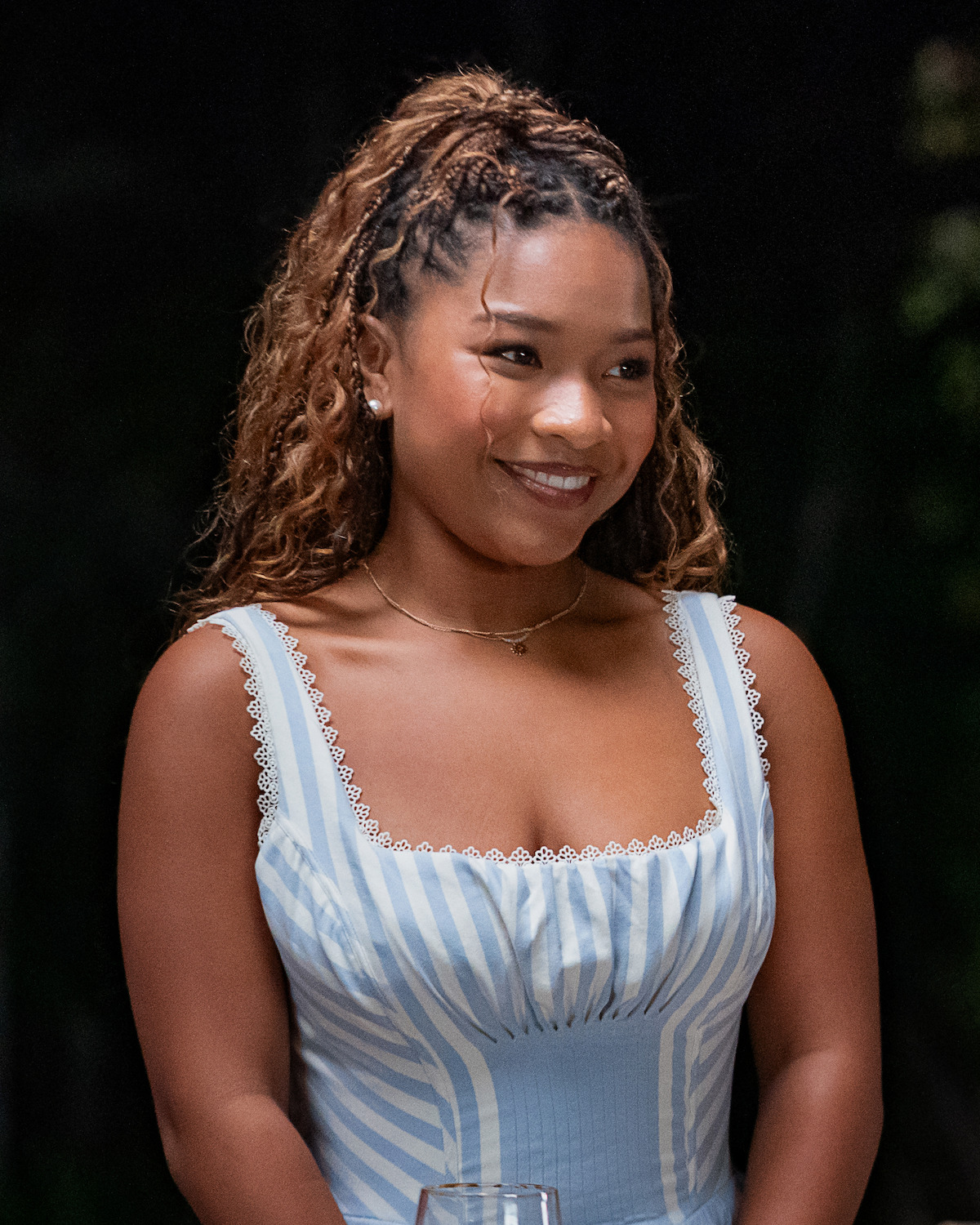 Young woman with curly hair styled half-up, wearing a blue and white striped dress with lace trim, smiling in an outdoor evening setting with a dark blurred background.