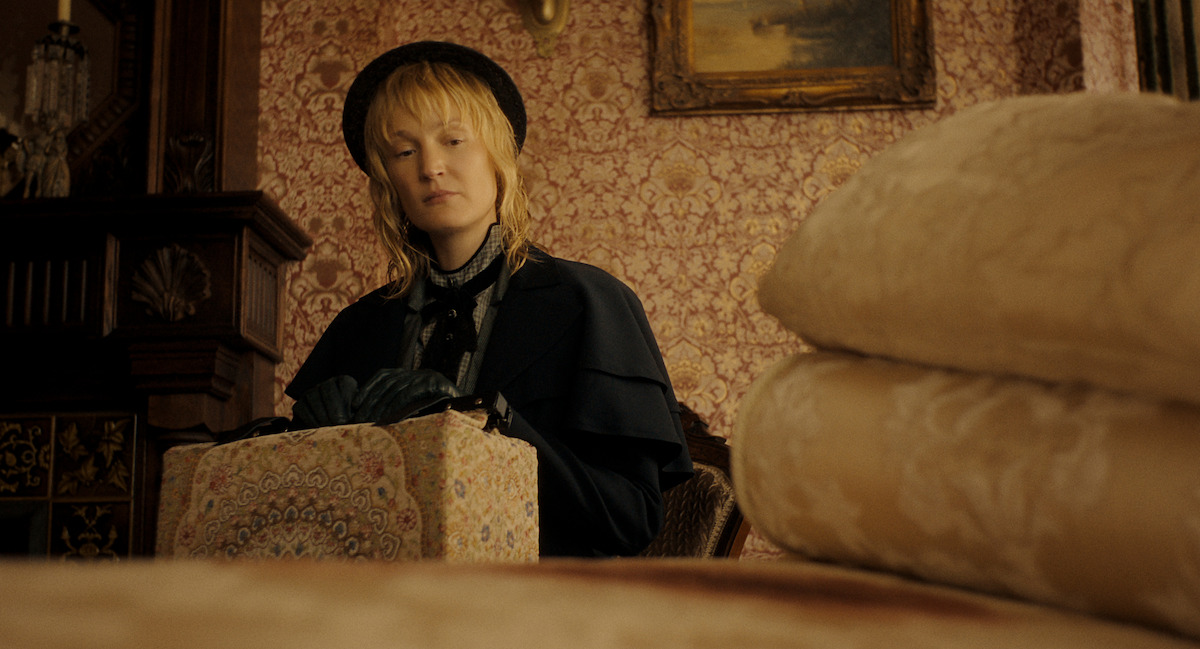 A woman in vintage clothing and gloves sits indoors, holding a patterned box. The room has ornate wallpaper, a painting, and folded linens in the foreground, suggesting a historical or period setting.