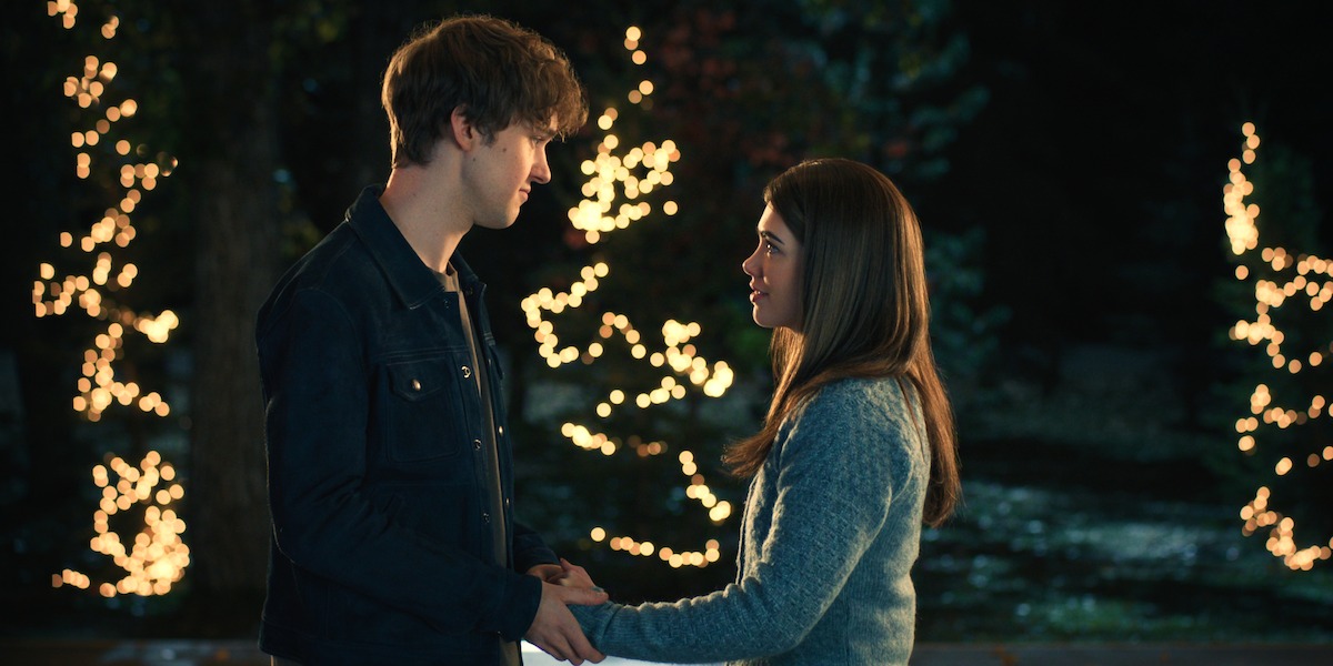 A young man and woman stand outdoors at night, holding hands and looking at each other, with trees decorated in glowing lights in the background.