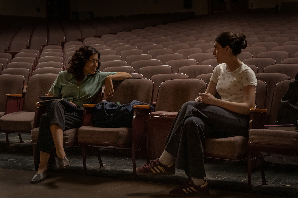 Two women sitting and talking in an empty theater auditorium, surrounded by rows of vacant seats, with bags on the chairs, suggesting a quiet discussion or meeting in a dimly lit, spacious environment.