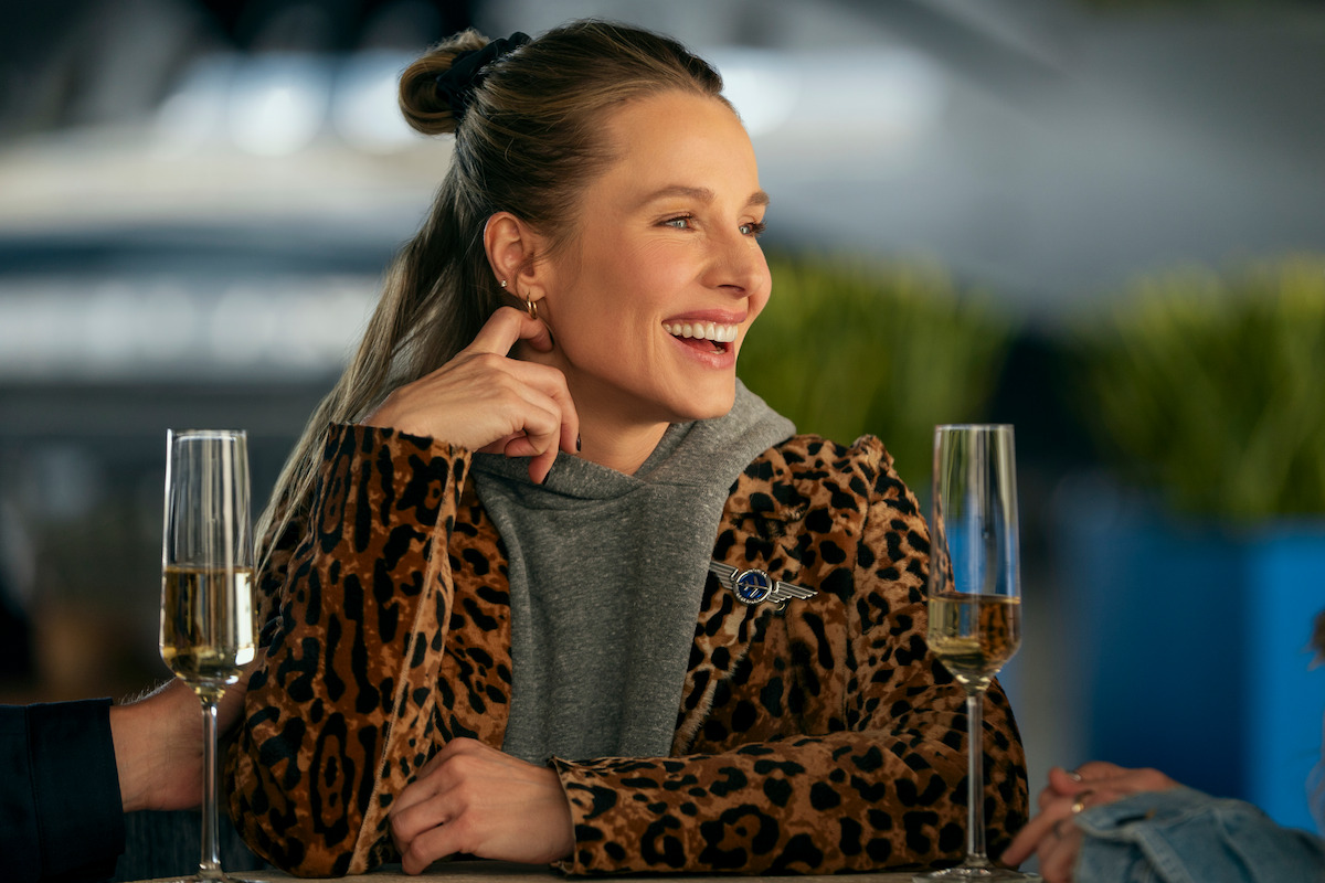 Woman in leopard print jacket sitting at a table, smiling and talking with others, two glasses of champagne in front of her, modern indoor setting with blurred background and plants.