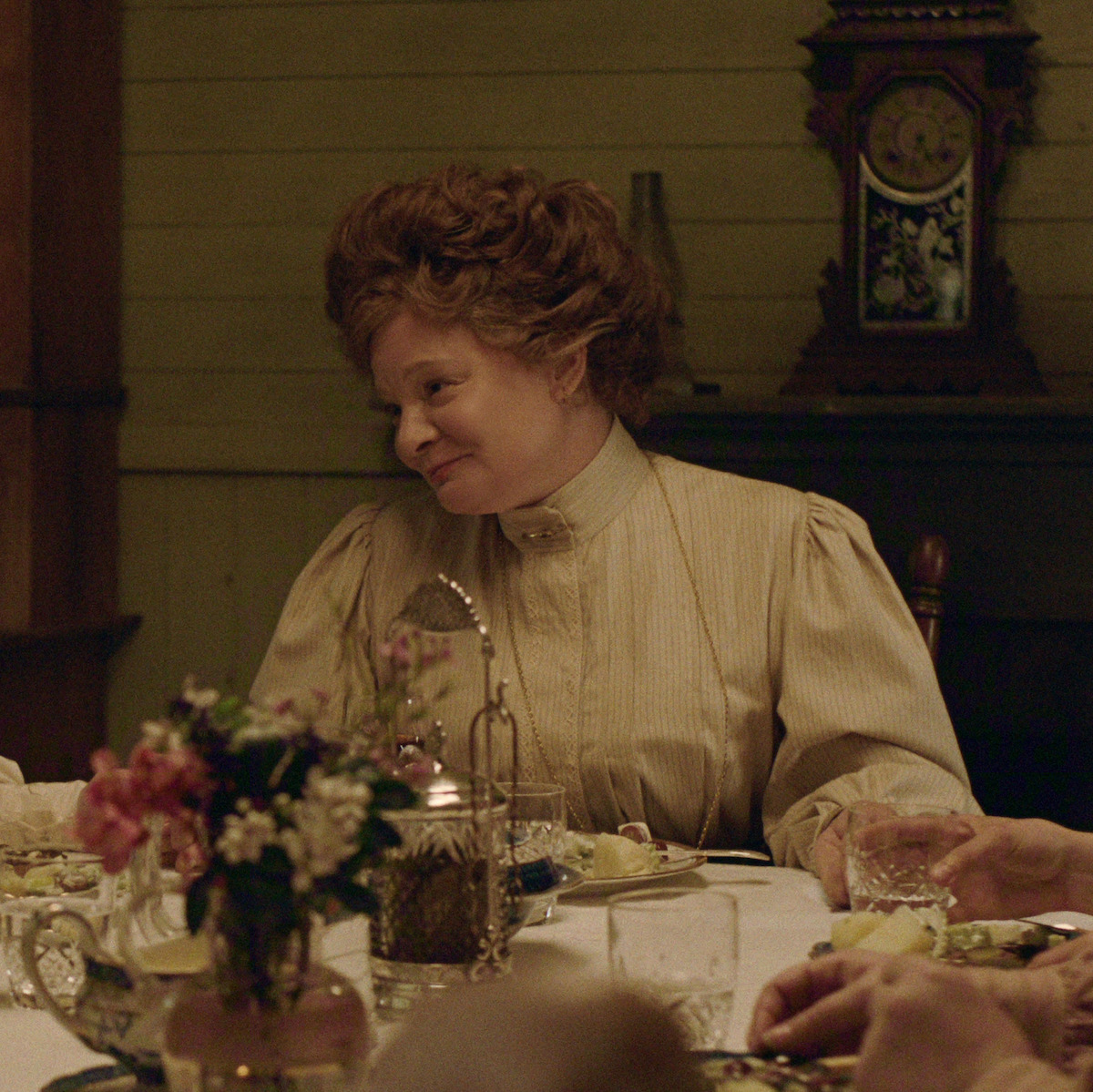 Martha Plimpton como Faye, com roupas de época, sentada a uma mesa de jantar posta com flores e louças, em uma sala antiga de iluminação acolhedora, com um relógio decorativo ao fundo.