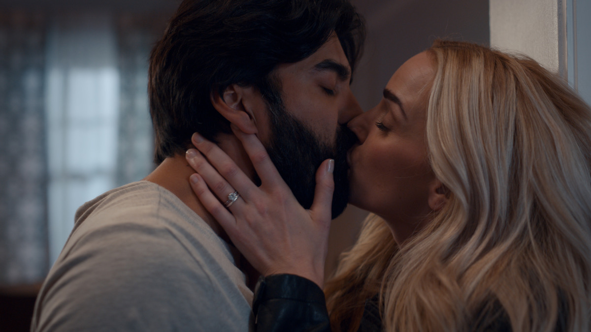 A man and woman share a passionate kiss indoors, with soft natural light from a window in the background, highlighting the woman's engagement ring and creating an intimate, romantic atmosphere.