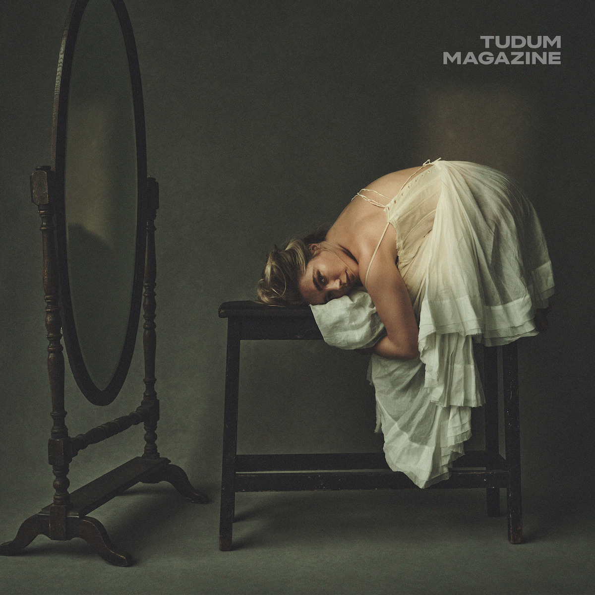 Woman in a vintage white dress curled on a dark wooden bench, head resting on cushion, near an antique standing mirror in a dimly lit studio with a moody, artistic atmosphere. Tudum Magazine logo in the corner.