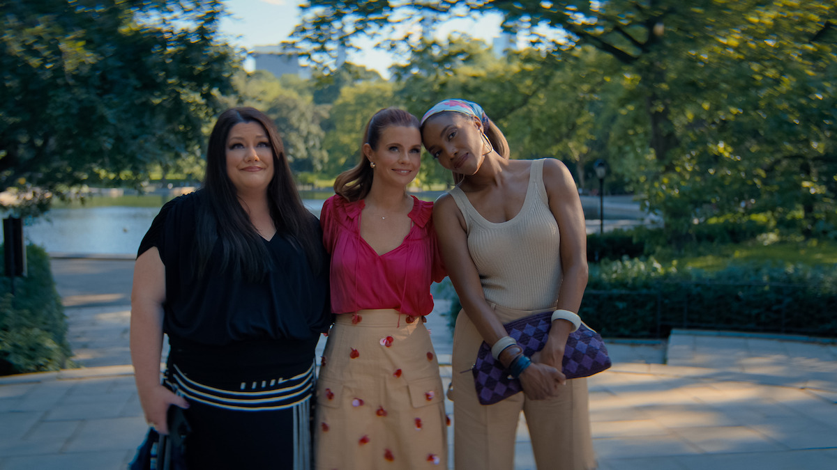 Three women standing closely together outdoors in a park with green trees and a pond in the background, dressed in casual stylish clothing, smiling and enjoying a sunny day.