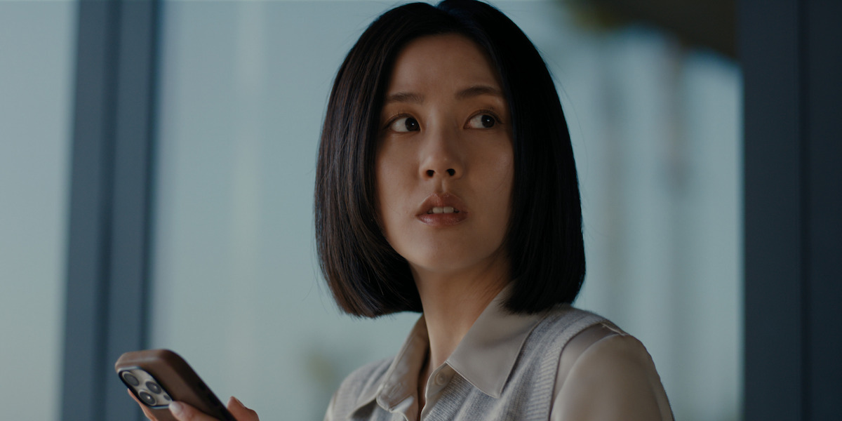 Woman with short dark hair holding a smartphone, indoors with large windows in the background, looking to her left with a serious or concerned expression.