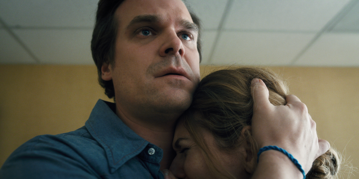A man in a blue shirt embraces a woman in a comforting hug in an indoor room with beige walls and a tiled ceiling.