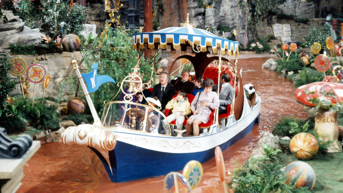 People riding in a decorated boat with a striped canopy on a chocolate river, surrounded by oversized candy and colorful whimsical scenery in a fantasy setting.