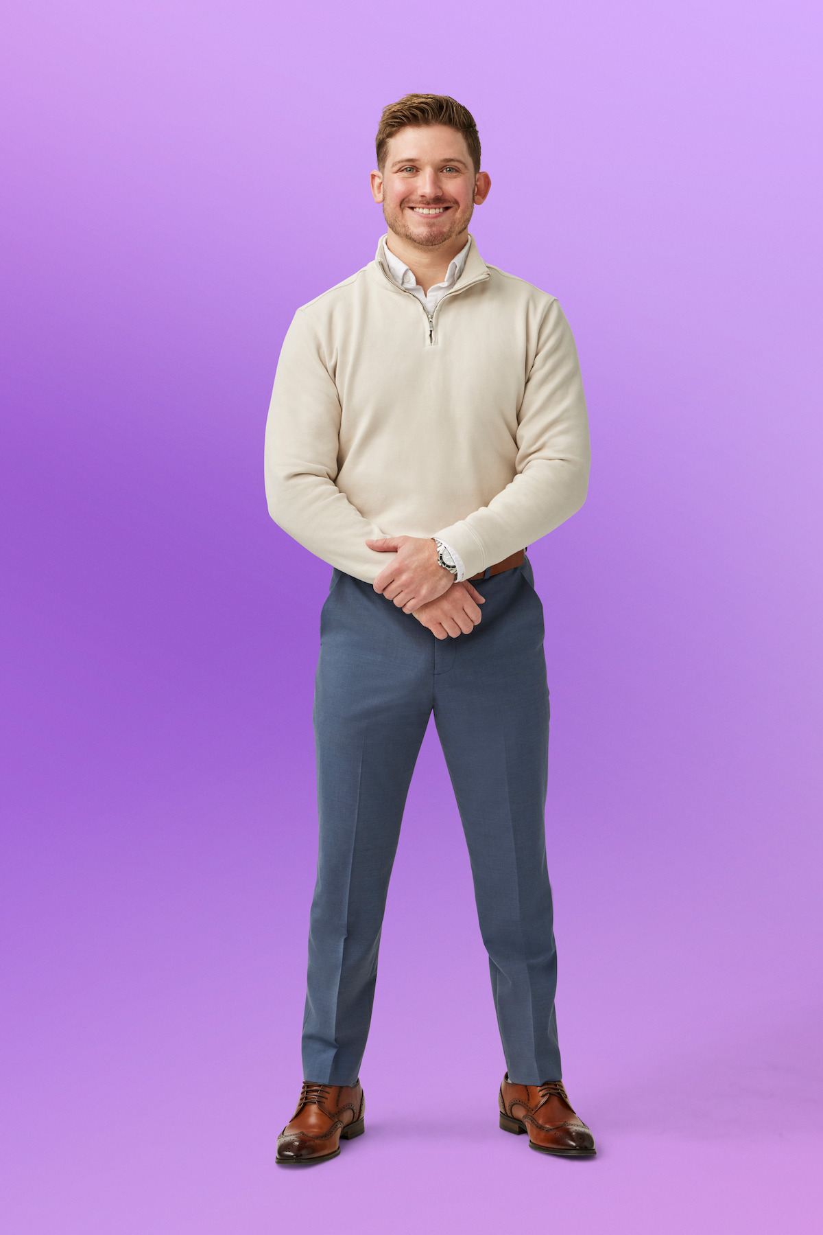 Chris in business casual attire with beige pullover, blue pants, and brown shoes stands confidently against a plain purple background in a studio setting.
