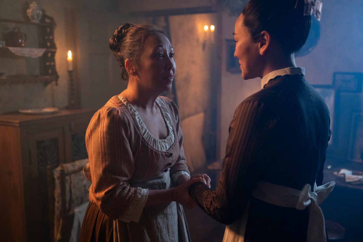 Two women in historical clothing stand close and hold hands in a dimly lit, candlelit room with rustic furniture, suggesting an emotional or intense conversation in a period drama setting.
