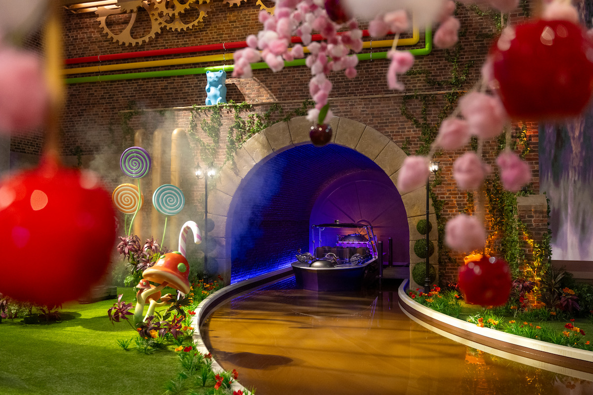 A whimsical indoor scene with a chocolate river, a boat entering a tunnel, oversized candy, lollipops, and cherry decorations, set in a colorful, fantasy factory environment with brick walls and playful gears.