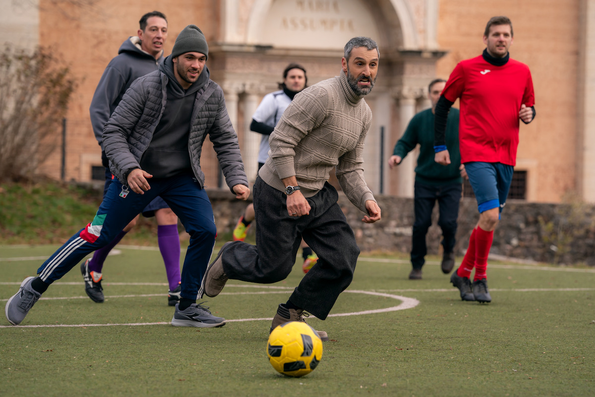 Group of men playing soccer on an outdoor field, with one man dribbling a yellow ball while others chase him. The setting is urban, with a historic building in the background and greenery along the field’s edges.