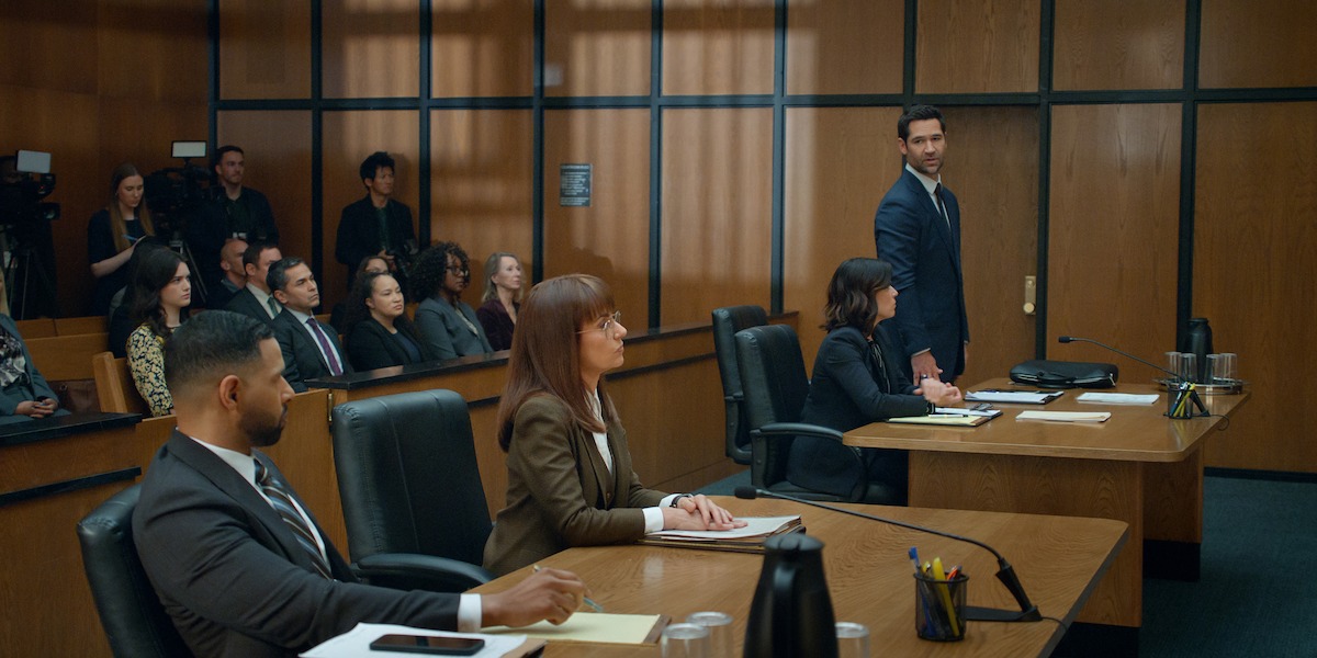 A courtroom scene with lawyers seated at tables, a man standing to speak, and people observing in the audience. The environment is formal with wood-paneled walls and typical courtroom furnishings.