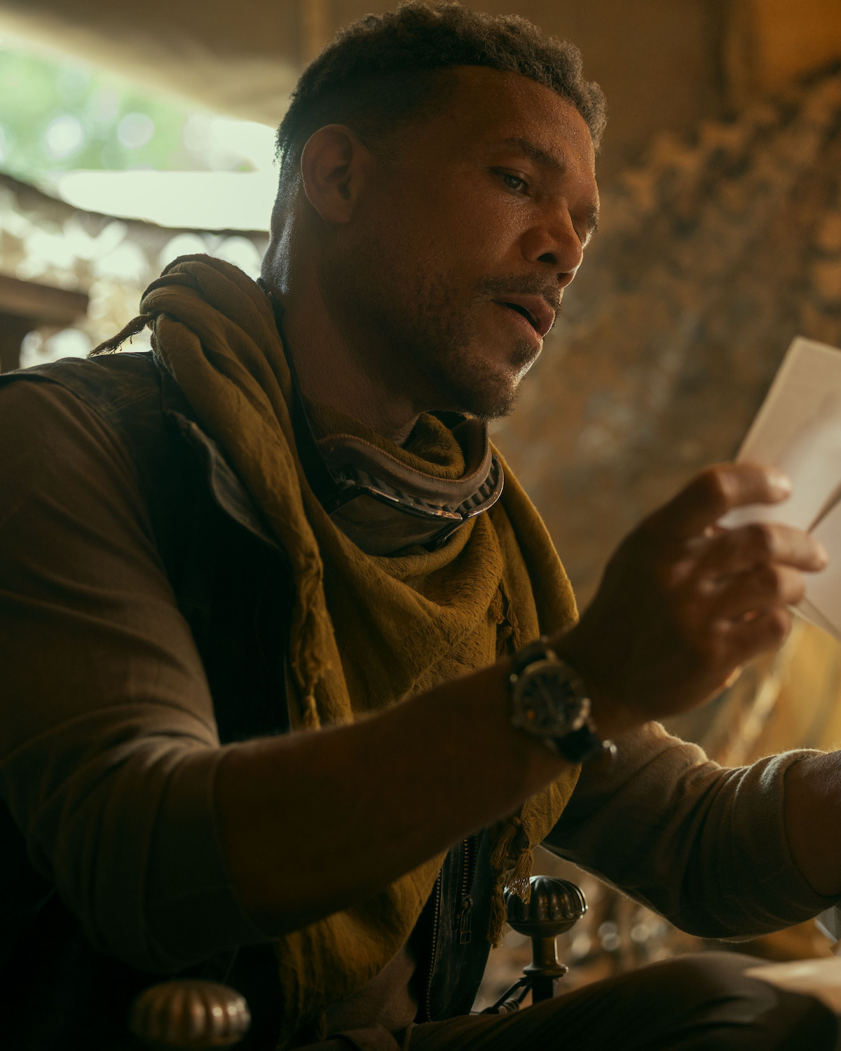 Jason Alan Carvell as Finch, in rugged outdoor clothing with a scarf and goggles examines a paper inside a tent-like structure with warm lighting, suggesting an adventurous or post-apocalyptic environment.