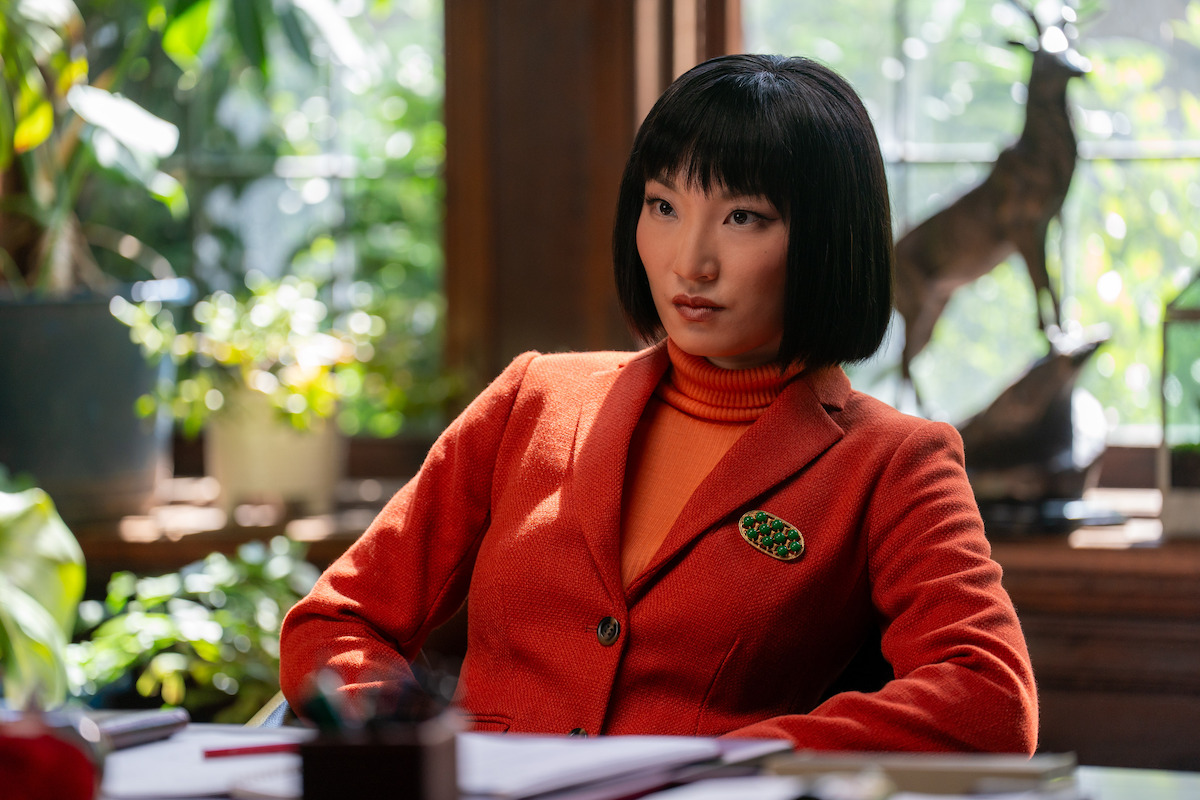 Woman in a red blazer sits at a desk in a sunlit, plant-filled office with wooden decor and a deer sculpture.