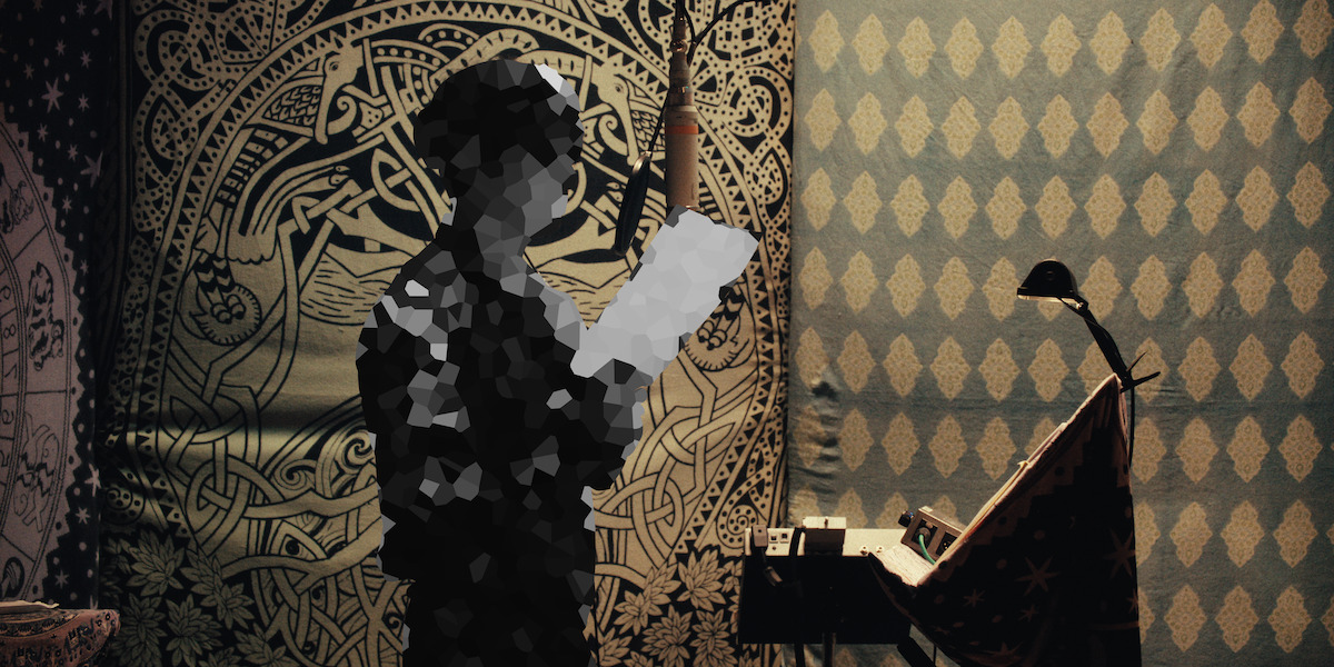 Pixelated person reading a paper in a decorated room with patterned tapestry on walls, a microphone overhead, and a lamp-lit desk with recording equipment, giving a creative and artistic studio environment.