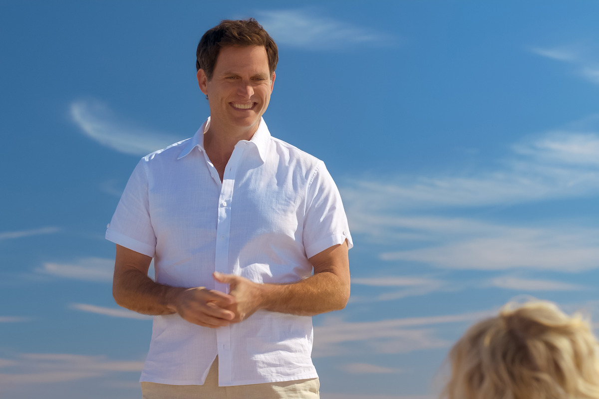 Smiling man in a white short-sleeve shirt standing outdoors under a bright blue sky with wispy clouds, talking to another person whose head is partially visible in the lower right foreground.