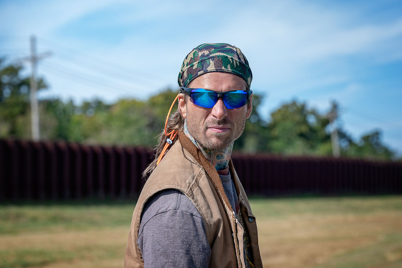 Gary Johnson (Glen Powell) undercover. He wears a camo hat, blue shades, and a brown vest.