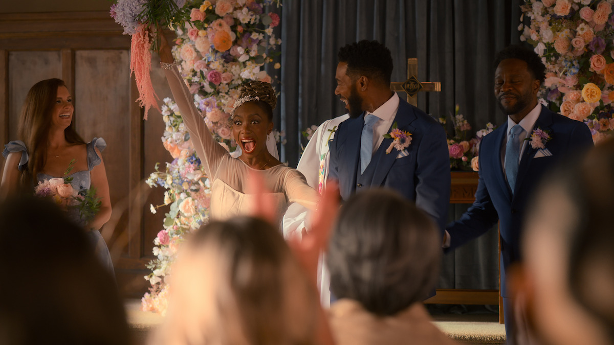 Bride and groom celebrate at indoor wedding ceremony with floral decorations, bridesmaid smiling on left, guests applauding in foreground, joyful atmosphere.
