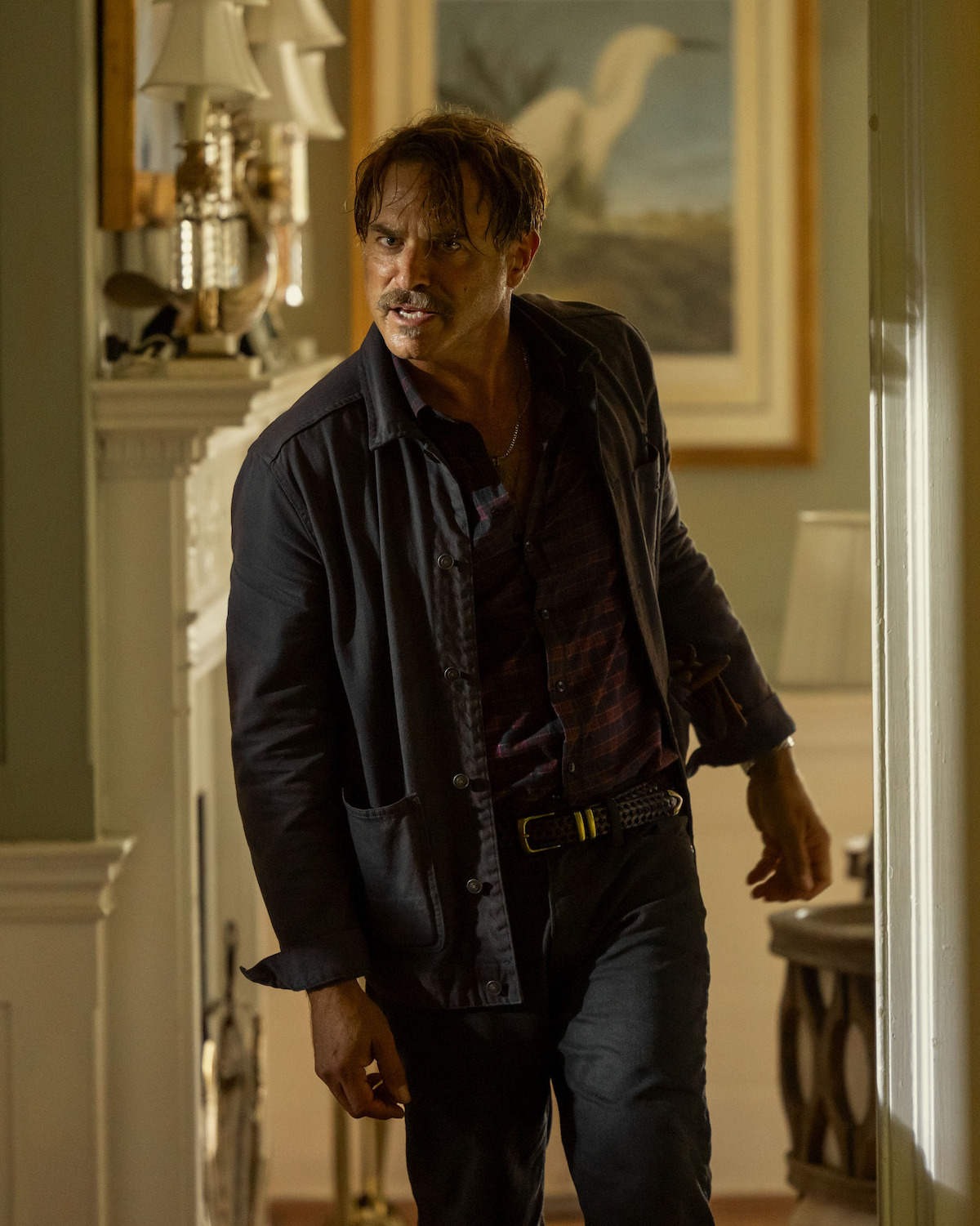 J. Anthony Crane as Chandler Groff, with a mustache, wearing a dark jacket and shirt, stands in a warmly lit, traditionally decorated room with framed art and lamps, appearing tense and alert as he looks toward the doorway.