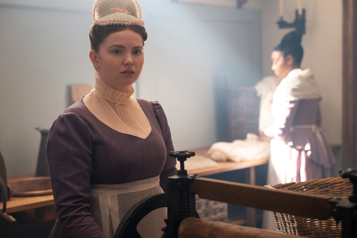Young woman in historical costume stands in a vintage laundry room with soft lighting, wearing a maid's outfit and cap, while another woman works in the background near baskets and wooden tables.