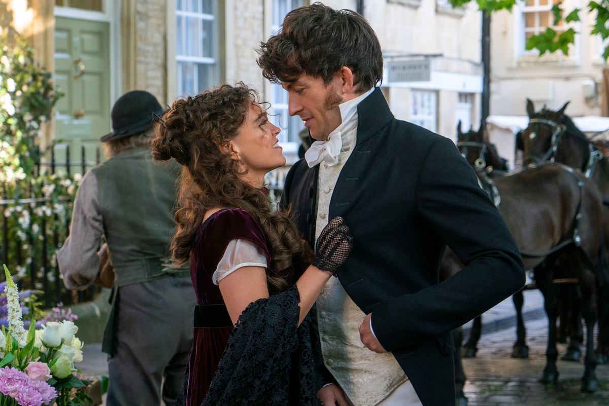 A woman and man in historical costumes gaze at each other on a cobblestone street with horses and carriages in the background, suggesting a romantic moment in a period setting.