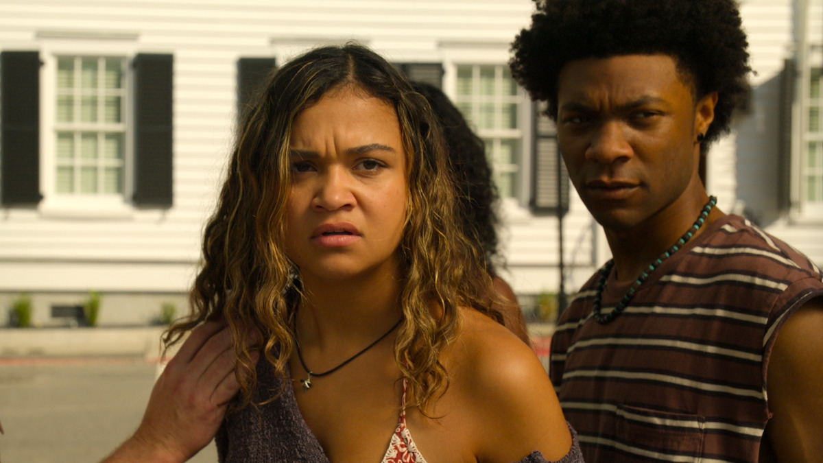 Two young adults stand close together outdoors in front of a white building with black window shutters, looking intently ahead with concerned expressions, suggesting a tense or dramatic situation.