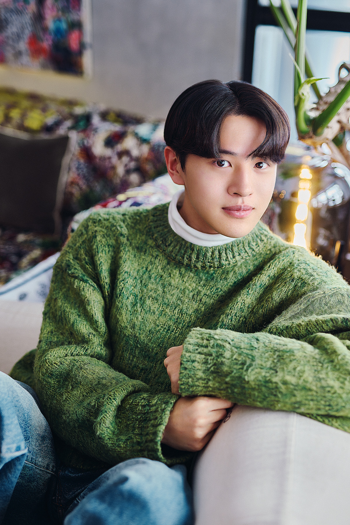 Young man with dark hair in a green sweater sits on a beige couch in a cozy, modern living room with plants and colorful decor, looking relaxed and content. Warm, inviting atmosphere with soft natural light.
