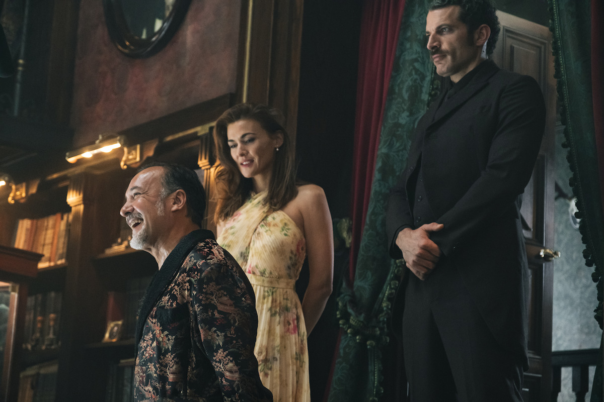 Three people in elegant attire stand in a richly decorated room with dark wood, bookshelves, and curtains, appearing to be engaged in conversation or a formal gathering.