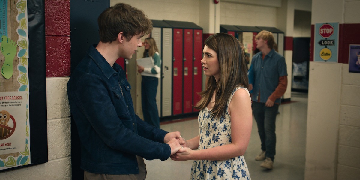 A teenage boy and girl hold hands in a high school hallway near lockers, facing each other and talking seriously. Other students and colorful posters are visible in the background, creating a typical school setting.