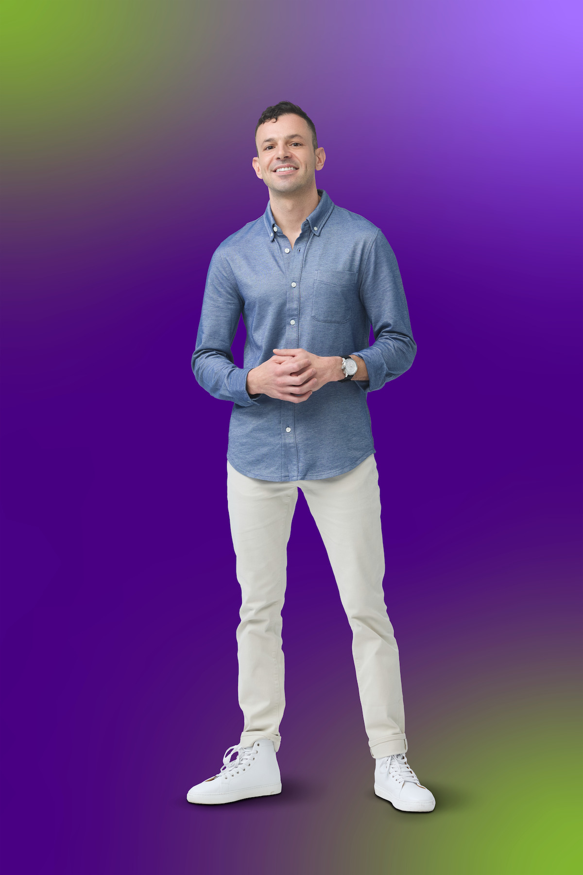 Man standing and smiling, wearing a blue shirt, light pants, and white sneakers, with hands clasped in front. Gradient purple, green, and yellow background, studio setting.