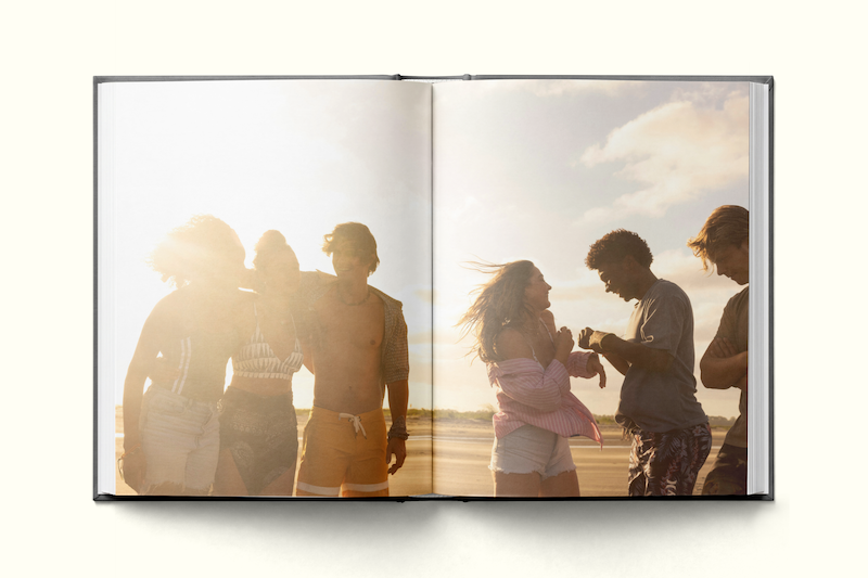 A group of young friends walking together on a sunny beach, laughing and enjoying themselves, pictured in a photo book with a bright sky and sandy background.