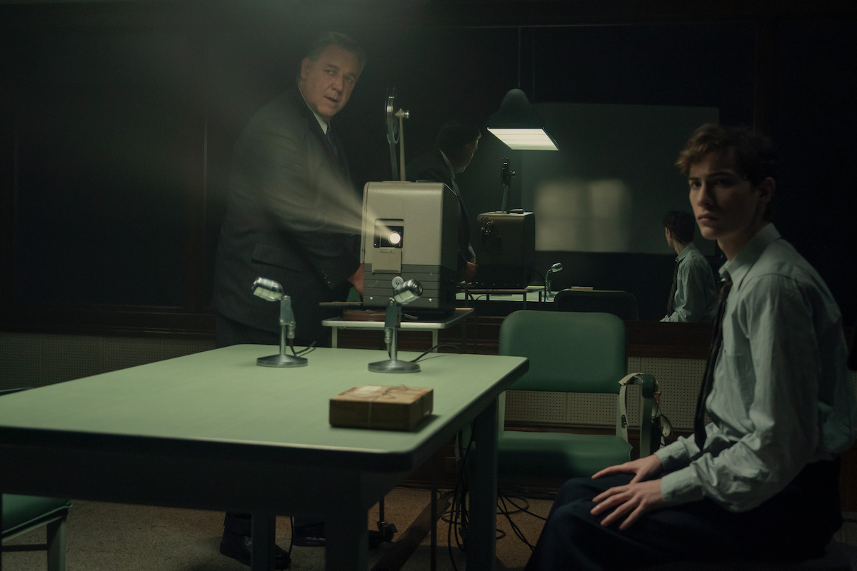 Two men in formal attire are in a dimly lit interrogation room with a green table, microphones, and a film projector. One man stands by the projector while the other sits, suggesting a tense interview or investigation setting.